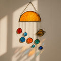 Three handcrafted stained glass suncatchers depicting the solar system and planets, featuring semi-circle, sun, and large sun designs – with vibrant colors like yellow, red, blue, orange, and green – hanging by metal chains in front of a sunlit window.