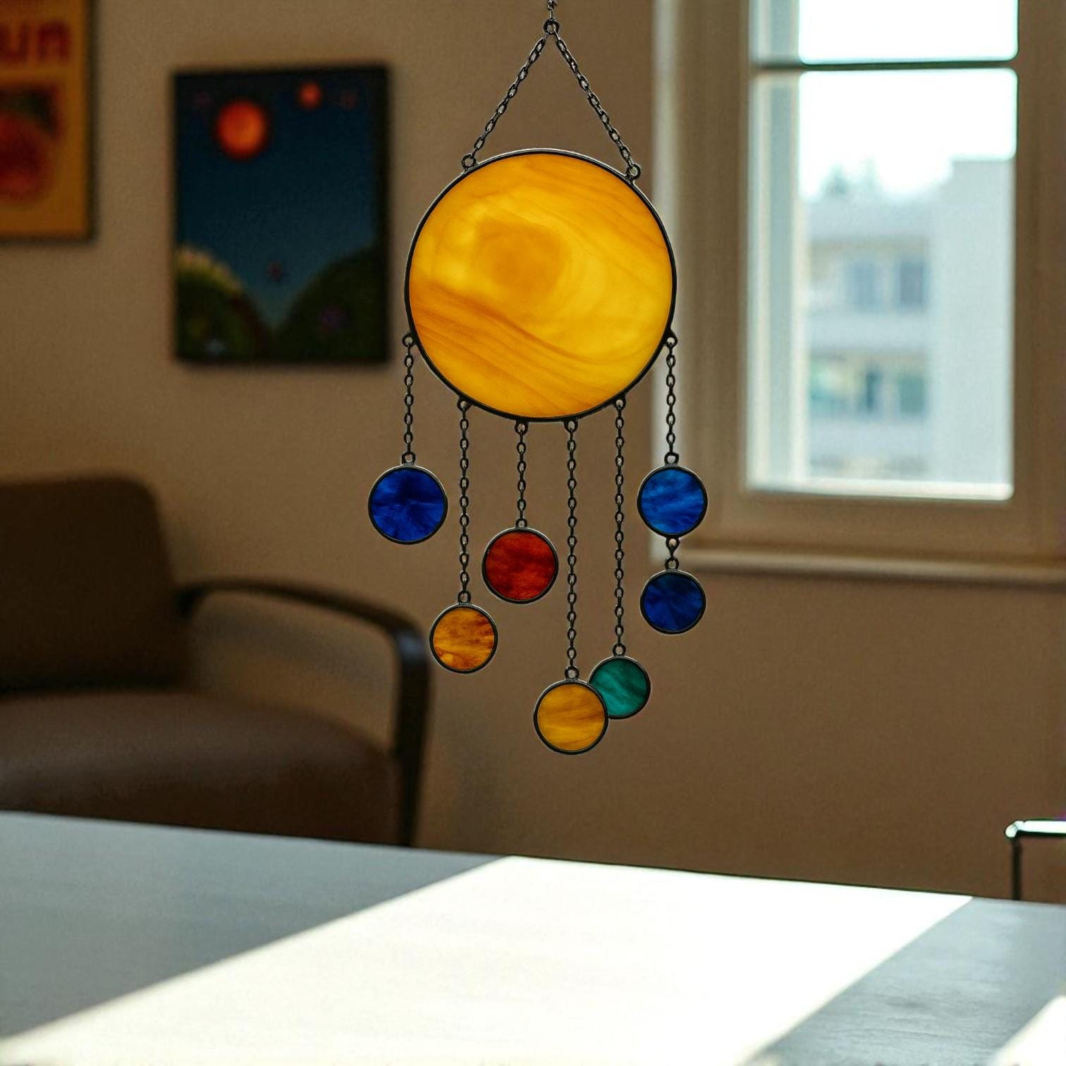 Three handcrafted stained glass suncatchers depicting the solar system and planets, featuring semi-circle, sun, and large sun designs – with vibrant colors like yellow, red, blue, orange, and green – hanging by metal chains in front of a sunlit window.