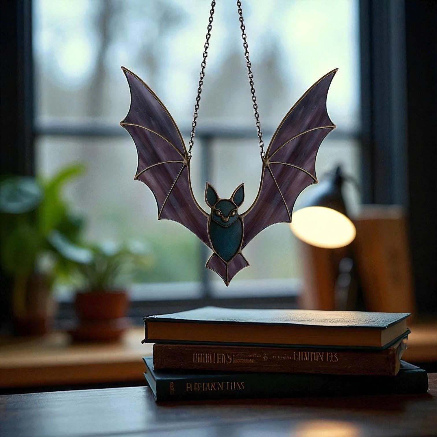 Stained glass bat suncatcher in purple glass hanging near books on a desk. Unique handmade gothic home decoration and witchy Halloween suncatcher.