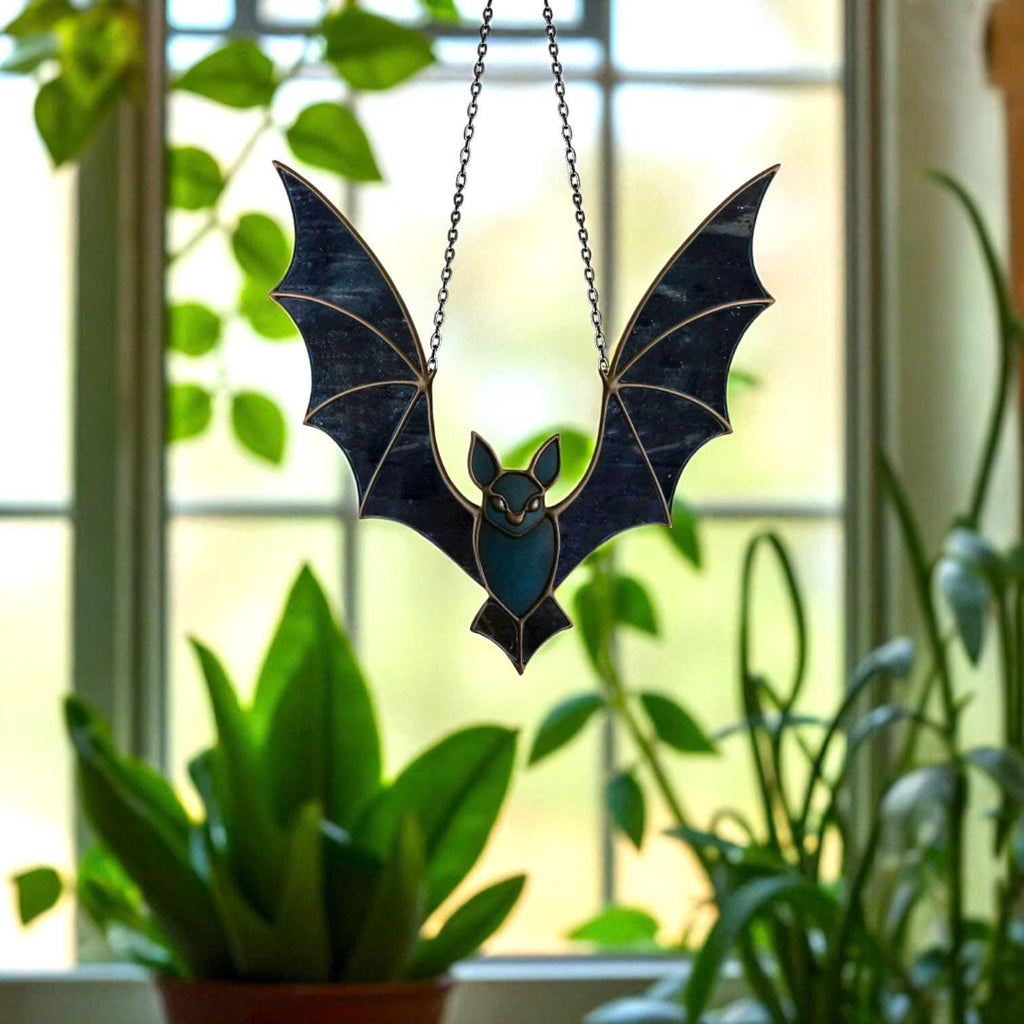 Black stained glass bat suncatcher hanging in front of a window with green plants in the background. Elegant gothic Halloween window hanging, handmade with real colored glass