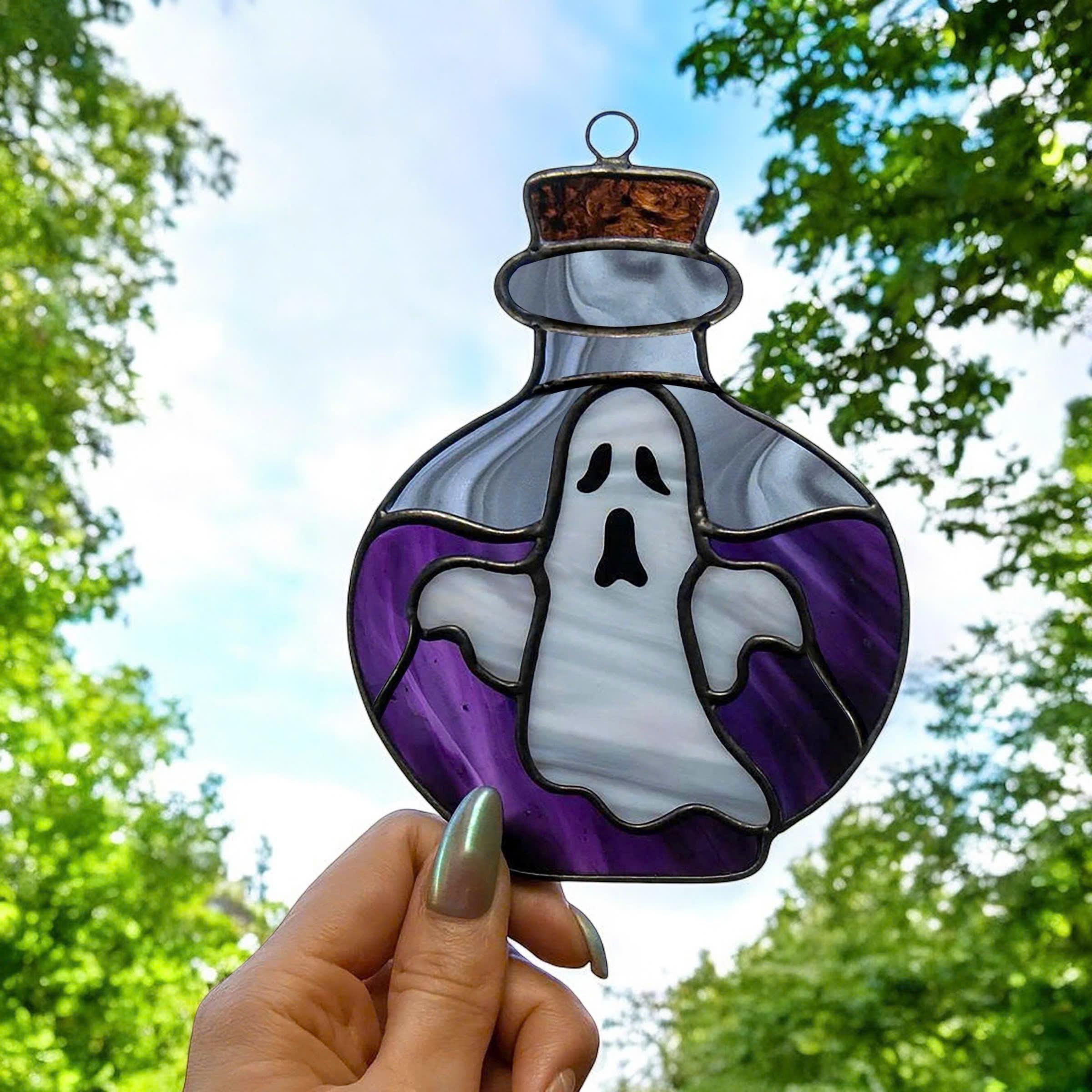 Handmade stained glass Halloween suncatcher shaped like a potion bottle with a white ghost inside, purple glass background, copper cork top, photographed outdoors against a blue sky and trees.