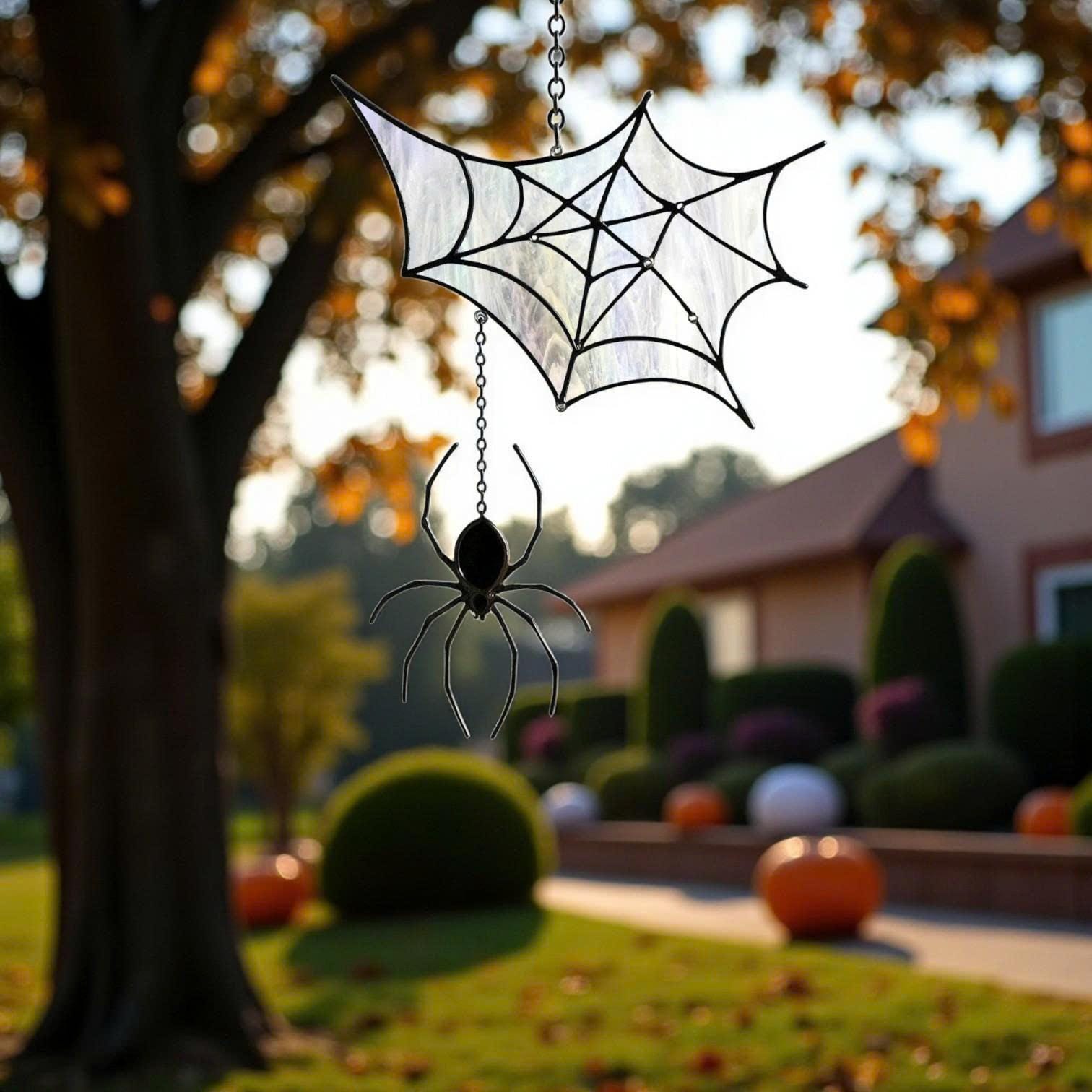 Handmade stained glass spider web suncatcher hanging on a chain from a tree branch, autumn leaves and a large house in the background, Halloween gothic decoration