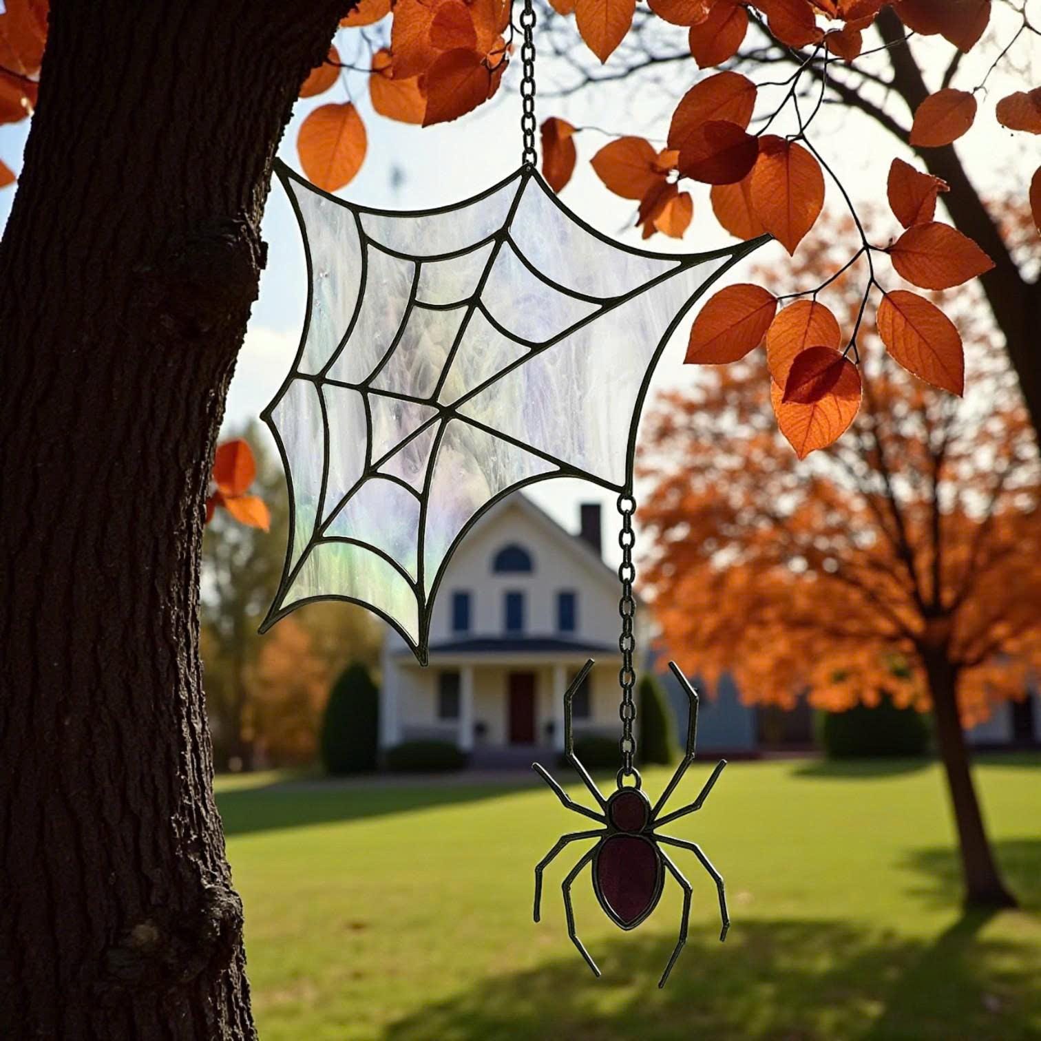 Close-up of stained glass spider web suncatcher with hanging spider, displayed outdoors with a white house and autumn trees in the background, handcrafted Halloween décor.