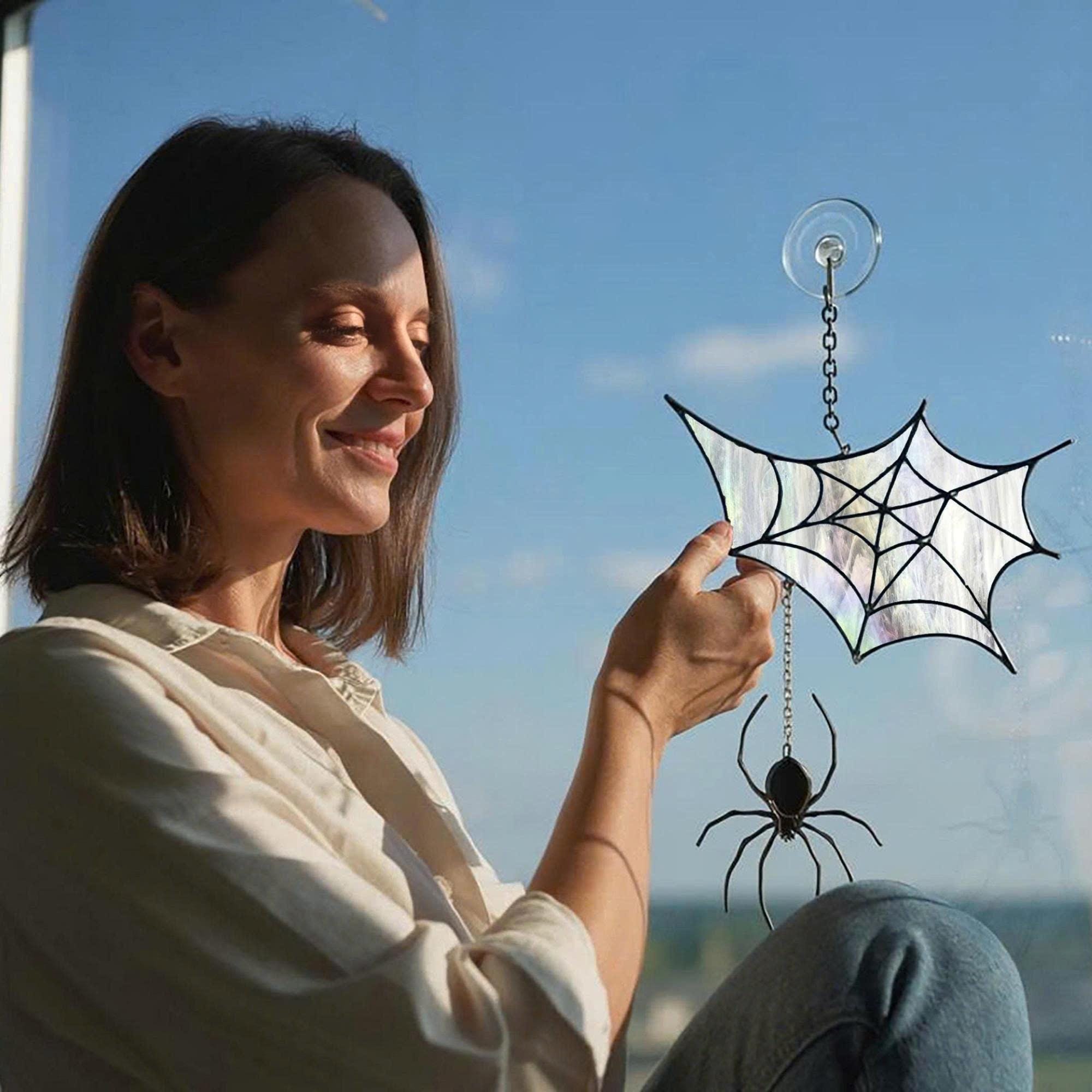 Smiling woman holding a stained glass spider web suncatcher with suction cup, sitting by a window on a sunny day, showing the product size and real glass detail.
