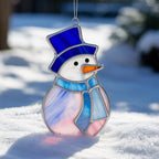 Stained glass snowman suncatcher (Style #9) hanging outdoors, with a fence and colorful blurred houses in the background.