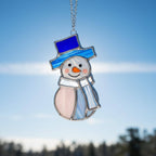 Stained glass snowman (Style #3) hanging on a snow-covered pine branch in a winter forest.