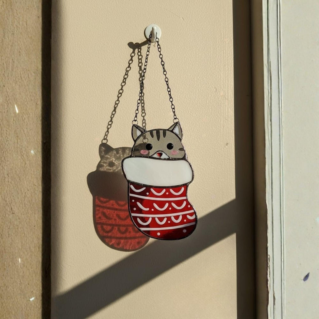 Handcrafted Christmas cat stained glass piece hanging on wall with bright sunlight reflection.