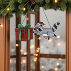 Raccoon stained glass ornament holding red and green gift, Christmas window hanging.