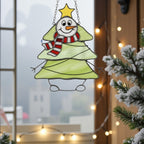 Snowman tree stained glass ornament glowing with winter sunlight on window sill.