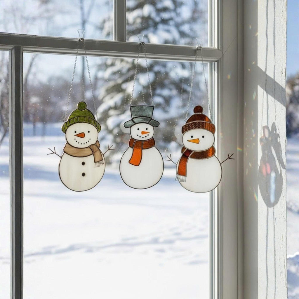 Handmade stained glass snowman ornaments hanging by window with snow background – winter Christmas home decor.