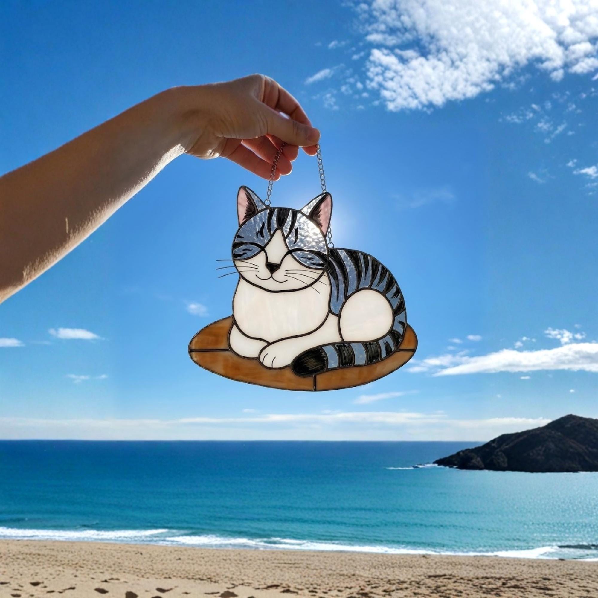 Grey striped cat suncatcher held against blue ocean background, summer gift idea.