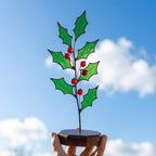 Hand holding stained glass holly branch against blue sky, showing transparent green glass and red berries.
