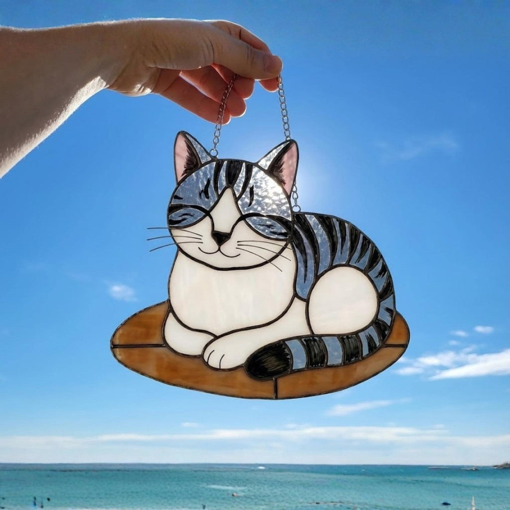 Hand holding grey striped cat suncatcher against blue sky, showing translucent glass texture.