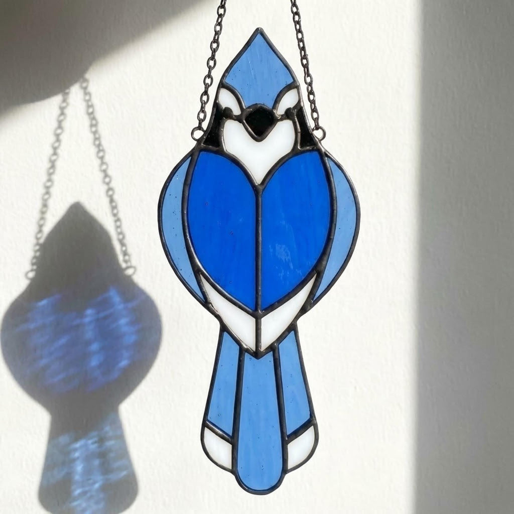 Stained glass Blue Jay bird suncatcher on branch hanging on window casting blue shadows, handmade realistic nature decor for birdwatchers by LuzyGlassArt.
