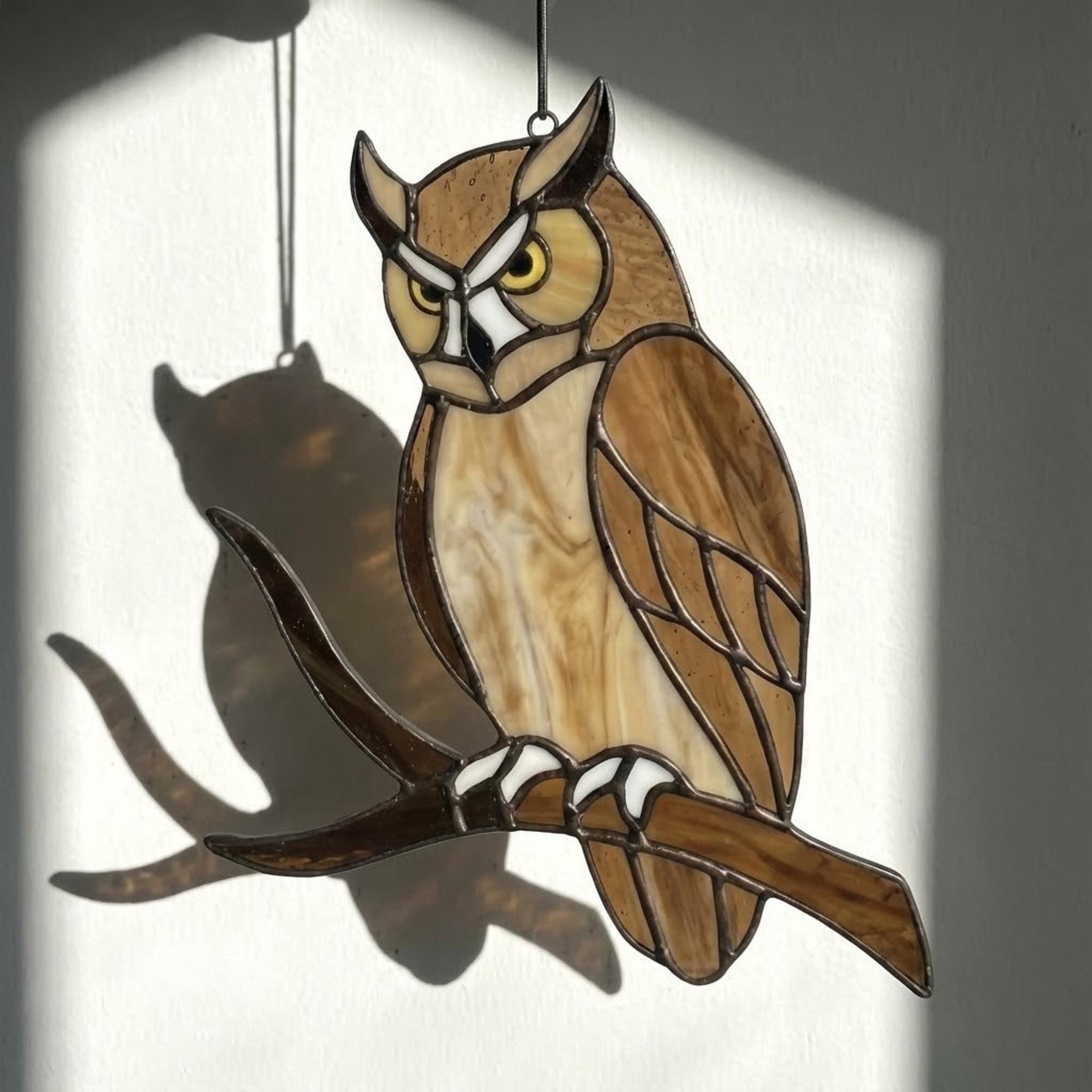 Handmade stained glass owl suncatcher in brown and beige tones, perched on a branch, casting warm amber shadows on a white wall, Tiffany style soldering detail.