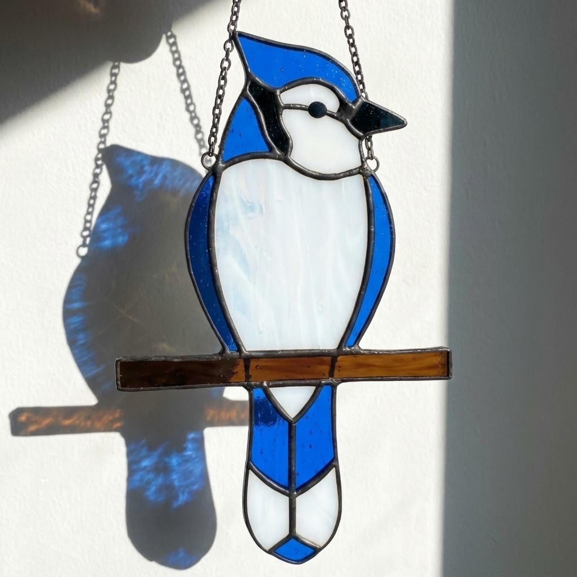 Stained glass Blue Jay bird suncatcher on branch hanging on window casting blue shadows, handmade realistic nature decor for birdwatchers by LuzyGlassArt.