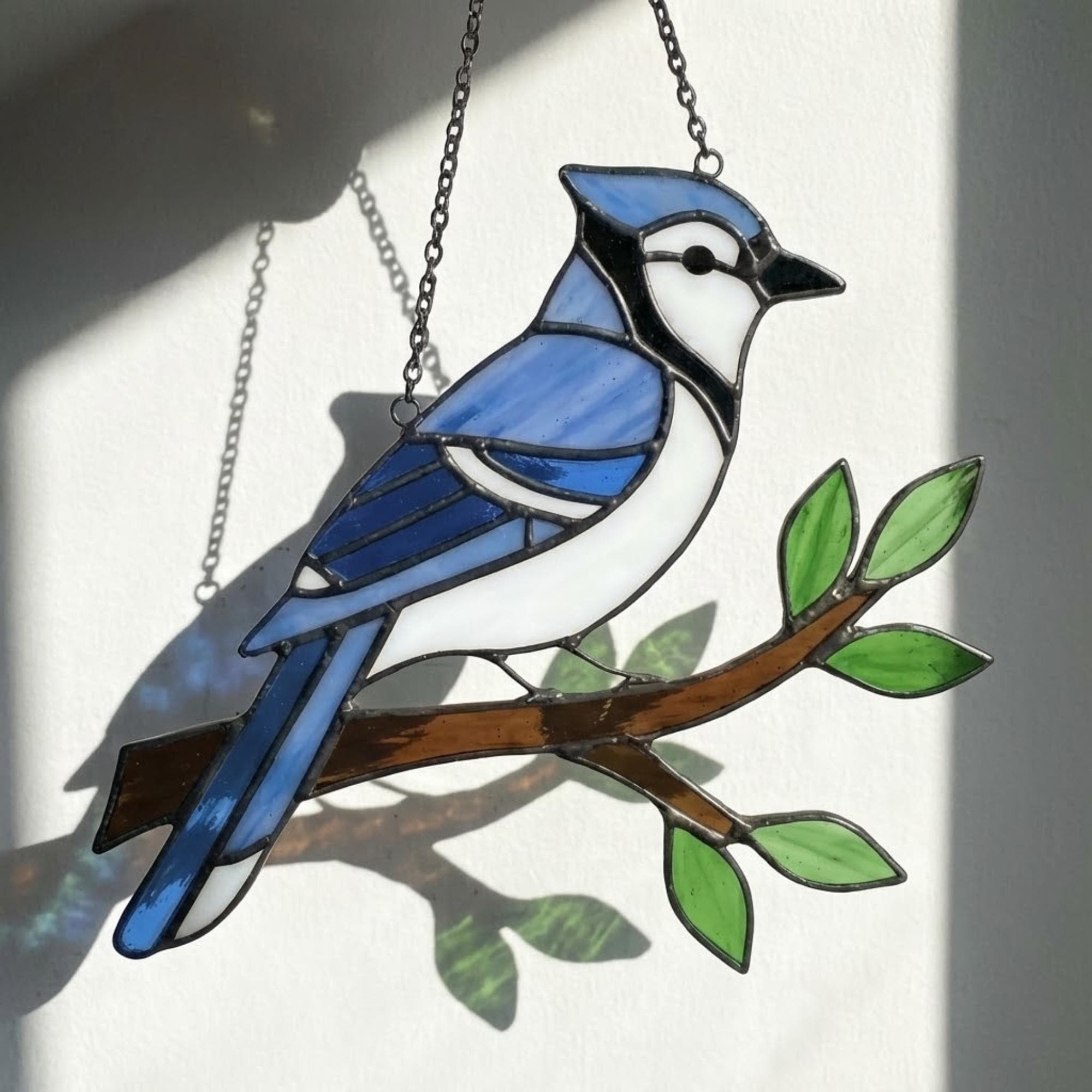 Stained glass Blue Jay bird suncatcher on branch hanging on window casting blue shadows, handmade realistic nature decor for birdwatchers by LuzyGlassArt.