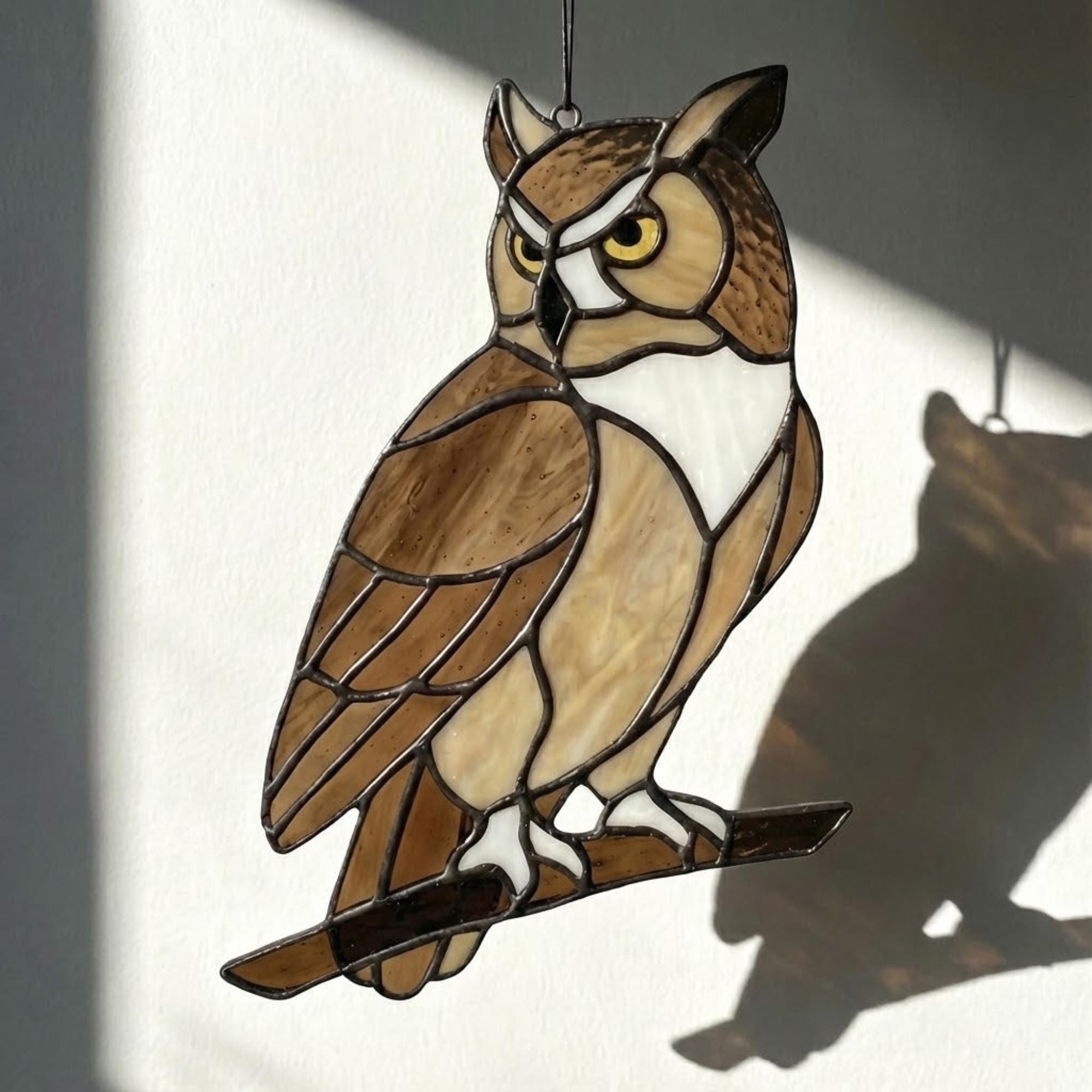 Handmade stained glass owl suncatcher in brown and beige tones, perched on a branch, casting warm amber shadows on a white wall, Tiffany style soldering detail.