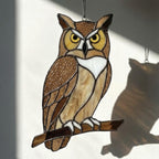 Handmade stained glass owl suncatcher in brown and beige tones, perched on a branch, casting warm amber shadows on a white wall, Tiffany style soldering detail.