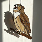 Handmade stained glass owl suncatcher in brown and beige tones, perched on a branch, casting warm amber shadows on a white wall, Tiffany style soldering detail.