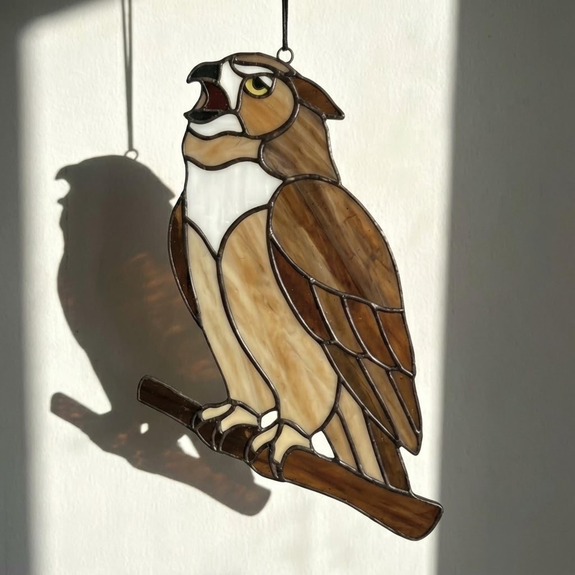 Handmade stained glass owl suncatcher in brown and beige tones, perched on a branch, casting warm amber shadows on a white wall, Tiffany style soldering detail.