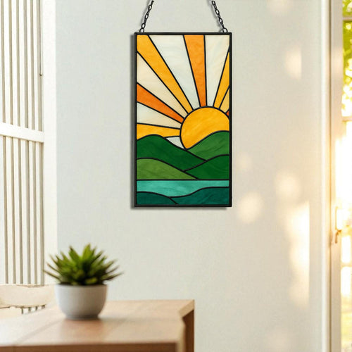 May include: Stained glass panel depicting a sunrise over a landscape. The artwork features a yellow sun with orange and white rays, green hills, and a blue lake. The panel is framed in black and hangs from a chain.