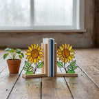 Yellow stained glass sunflower bookends with green leaves on a natural wood base holding books, catching spring sunlight on a desk.