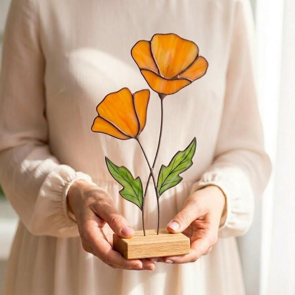 Handmade stained glass poppy flower sculpture with vibrant red petals and hand-painted veins, mounted on a solid oak wood base, casting a colorful warm glow on a sunny wooden table.