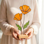 Handmade stained glass poppy flower sculpture with vibrant red petals and hand-painted veins, mounted on a solid oak wood base, casting a colorful warm glow on a sunny wooden table.