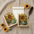 Yellow stained glass sunflower bookends with green leaves on a natural wood base holding books, catching spring sunlight on a desk.