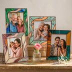 A collection of 6 different handmade stained glass photo frames in teal, amber, and pastel colors arranged on a sunlit wooden table.